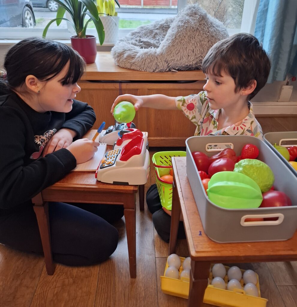 Children taking part in shop role play with a toy cash register and play food, supporting learning through play in home education.