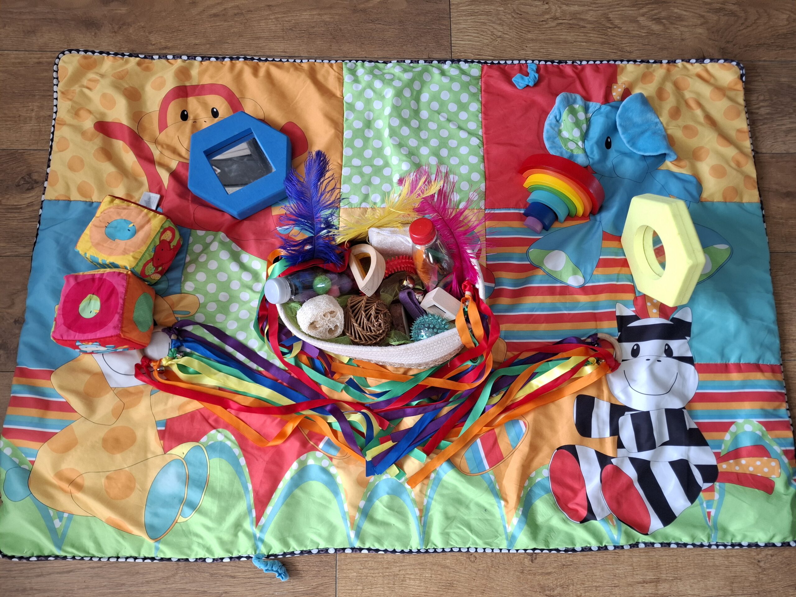 Baby area for younger party guests, with a colourful play mat, sensory basket, ribbon rings, feathers, soft blocks, mirrors and a wooden rainbow stacker