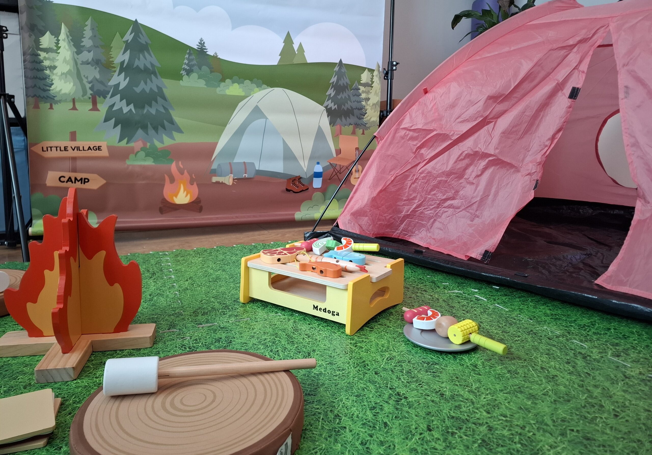 Camping site role play area for a children's party, with a pink play tent, wooden campfire, toy barbecue with play food, marshmallow toasting set and grass mat