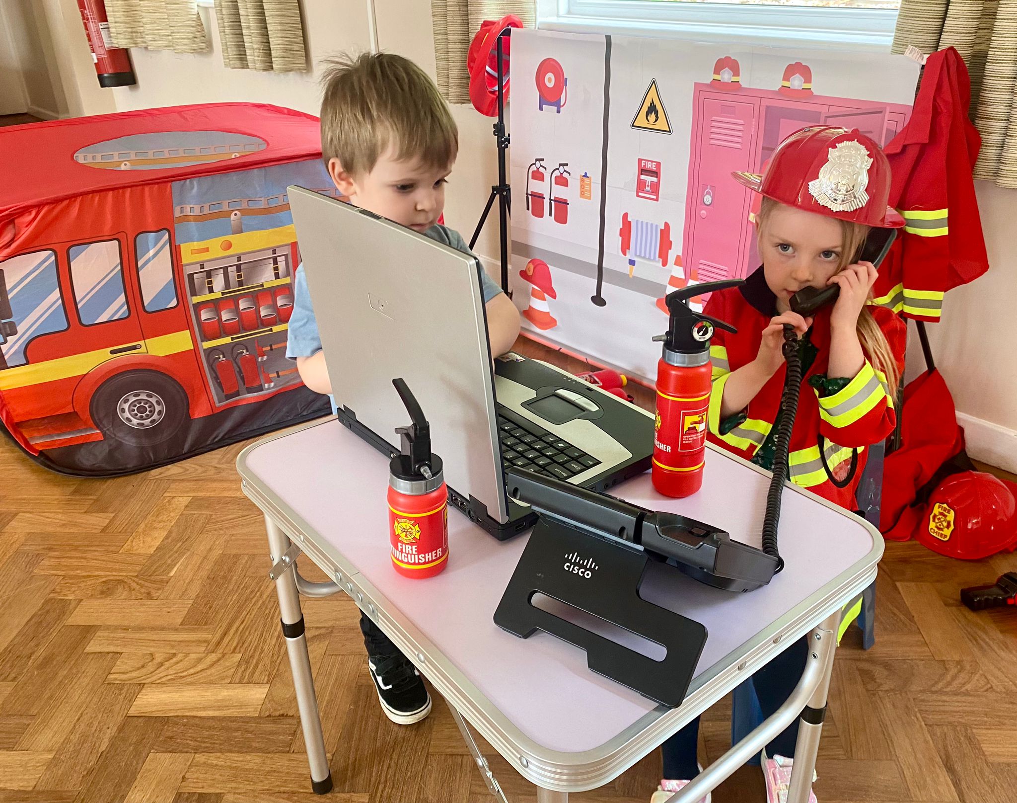 Two children playing fire station role play at a party, one in a firefighter helmet and jacket answering the phone, with a fire engine play tent